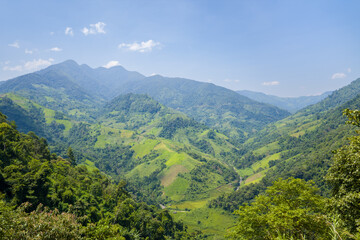 Sweeping view of a lush green valley surrounded by rolling forested mountains under a clear blue sky in northern Vietnam. Sunlight illuminates the layered hills and vibrant foliage, creating a sense