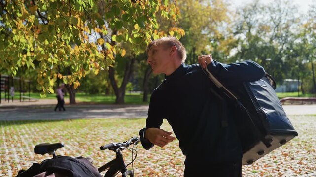 white delivery rider with bag bike, smiling and adjusting shoulder strap under golden trees, bicycle parked on path scattered with leaves, checking phone and giving thumbs up, sunlight warm, casual
