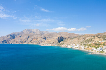 Fototapeta premium Wide view of Tsoutsouros bay in Crete with vivid blue water, a coastal village, and rugged sunlit mountains stretching beneath a bright, cloud-dotted sky.