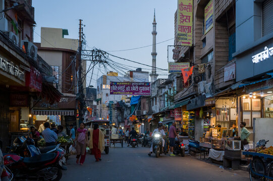 Lucknow, India - March 16, 2024: The image shows an urban street at dusk with pedestrians, motorcycles, illuminated shops, visible signage in Hindi, and a mosque minaret in the background.