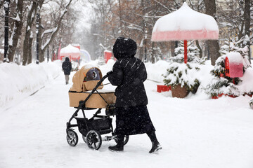 Woman with a baby stroller walking in winter park. Snow weather, concept of motherhood, single mom with pram