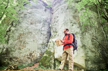 Bearded tourist man studies map while stands in front of large rock in dense forest. Traveler with grey backpack and glasses, planning outdoor adventure, hiking or climbing route.