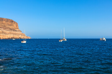 Boats sailing on deep blue ocean waters near a prominent coastal cliff face. Wide view of sea showing several vessels positioned offshore from a large, stratified rock formation