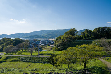 A peaceful rural village nestles among lush green hills and scattered trees under a clear blue sky near Hara, with distant mountains and water visible in the background. Crisp sunlight highlights the