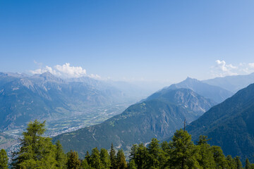Fototapeta premium Sweeping view over the Rhone Valley in Switzerland, featuring layers of rugged mountains, green forests, and a clear blue sky with soft clouds creating a tranquil summer atmosphere.