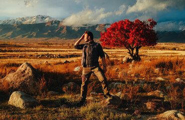 Heroic-looking man adjusting hair with one foot on rock in steppe with red tree and mountains