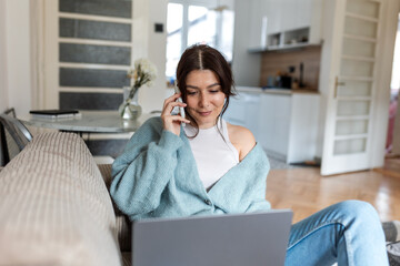 Focused young woman using laptop and talking on mobile phone at home. Female professional in blue cardigan multitasking in a bright modern apartment. Remote work and telecommunications concept.
