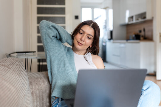 Young woman stretching her neck while working on a laptop at home. Female freelancer feeling tired or taking a break from remote work in a cozy living room. Concept of neck pain or office fatigue.