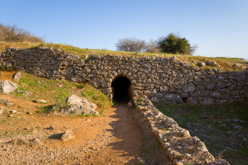 Fototapeta premium A weathered stone tunnel leads into the hillside at Palamidi Fortress in Nafplio, Greece. Sunlight highlights the rough masonry, dry earth, and sparse vegetation, creating a sense of mystery and