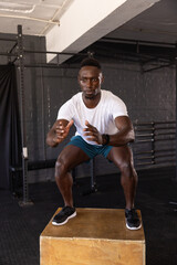 Naklejka premium African American man doing plyometric box jump at gym with wooden box, squat rack and smartwatch