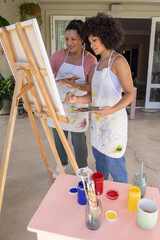 Obraz premium Diverse female friends painting at covered patio on wooden easel with paint jars and brushes