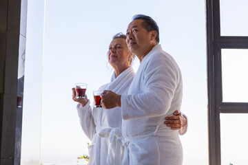 Diverse senior couple wearing bathrobes, holding glass tea cups at balcony door gazing horizon