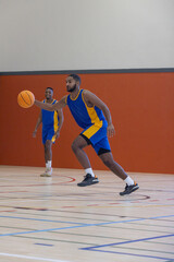 Naklejka premium African american male teammates dribbling basketball on court in uniforms, copy space