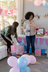 Fototapeta premium Diverse female friends preparing drinks near gift bags, balloons in living room