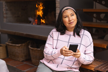 Naklejka premium Woman in her 40s sitting by fireplace holding smartphone near wicker baskets, gazing, copy space