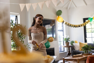 Fototapeta premium Asian woman wearing dress holding gold party horn at cake table, balloons in living room