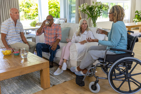 Senior friends chatting around coffee table in living room, snacking chips, sipping water