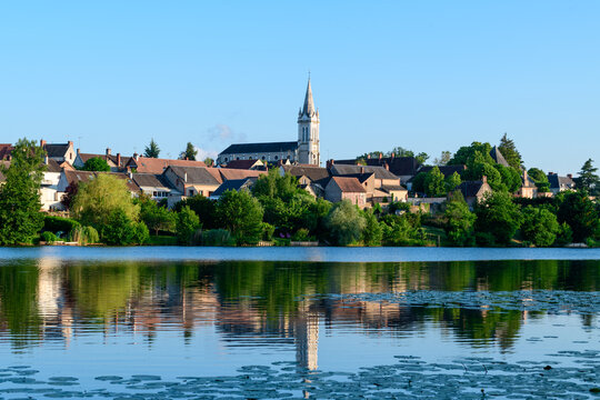 Warm sunlight bathes the rooftops and tall church spire of Dampierre en Burly, their forms mirrored in the still waters of a pond bordered by lush summer foliage.