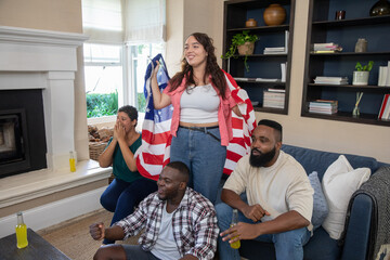 Naklejka premium Diverse friends cheering and draping american flag, holding glass bottles in cozy living room