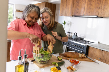 Diverse senior friends tossing salad at kitchen island with bowl, wooden servers, vegetables © wavebreak3