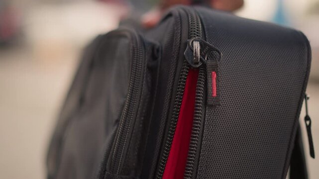 closeup black bag zipper red lining. tight shallow depth of field highlights textured synthetic fabric and metal slider as hand pulls zipper on urban sidewalk blurred cars and pedestrians suggest