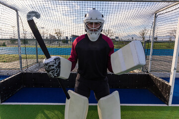 Obraz premium Field hockey goalkeeper is standing before goal cage on turf with stick wearing protective gear