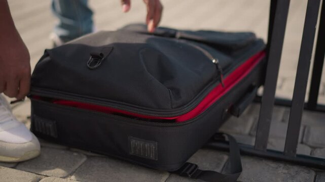 black man unzipping redlined equipment case on sidewalk, hands pulling heavy zipper beside metal fence, white sneakers visible, careful reveal of padded interior, musician preparing portable audio