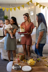 Fototapeta premium Diverse female friends celebrating in living room holding cake with lit candles, cradling baby