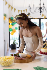 Fototapeta premium African american woman wearing strapless dress arranging buffet at home with charcuterie, cake
