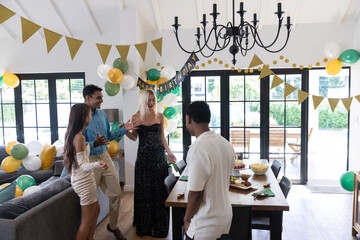 Diverse friends gathering around wooden dining table by sofa and glass doors, birthday cake