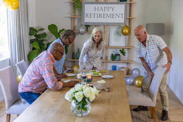 Naklejka premium Diverse senior coworkers preparing retirement party table in dining room with cupcakes, balloons