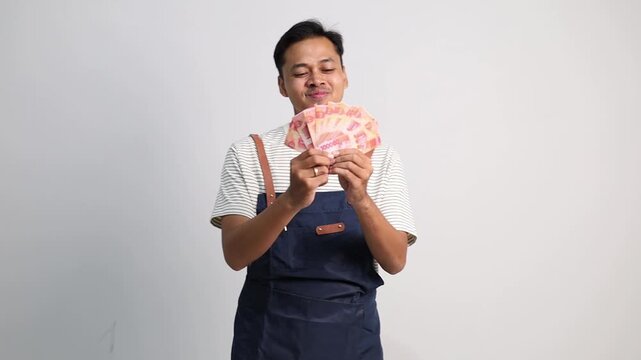 A young man wearing an apron carries several rupiah banknotes, with a happy and relieved expression of having received a profit from his business or salary.