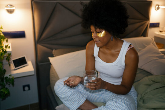 Woman sitting on bed taking pills with glass of water in bedroom