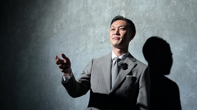 グレー背景でピンマイクをつけて話すスーツの日本人男性