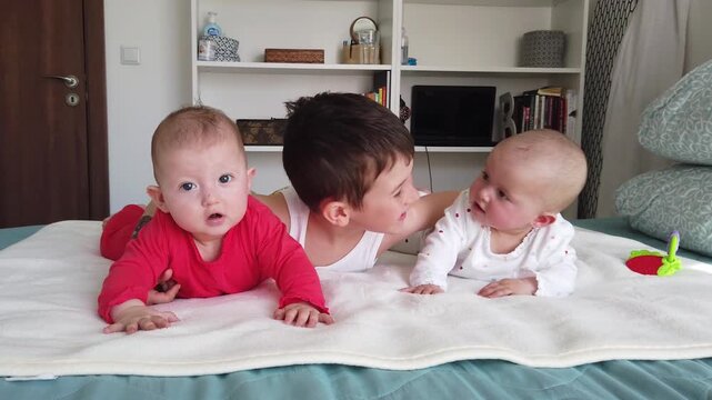 Joyful older brother plays with his cute twin baby siblings on a bed at home. A heartwarming scene of family love, care, and togetherness. Happy childhood and sibling bonding concept.