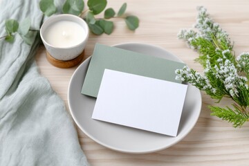 card mockup on a plate, with christmas decorations around it, a candle, and a green color theme, on a light wooden table background