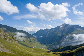 Expansive view of a green alpine valley framed by steep rocky mountains, scattered clouds, and a winding trail under a vibrant blue sky.