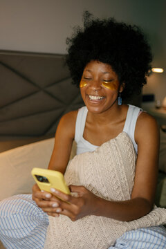 Laughing woman with eye patches using smartphone in bed in the morning