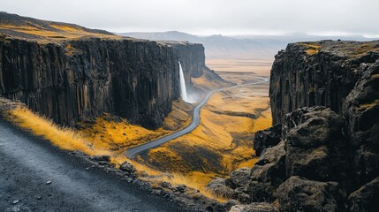 Obraz premium Isolated black road in iceland with tall rock cliffs on sides and yellow grass leading to distant waterfall.