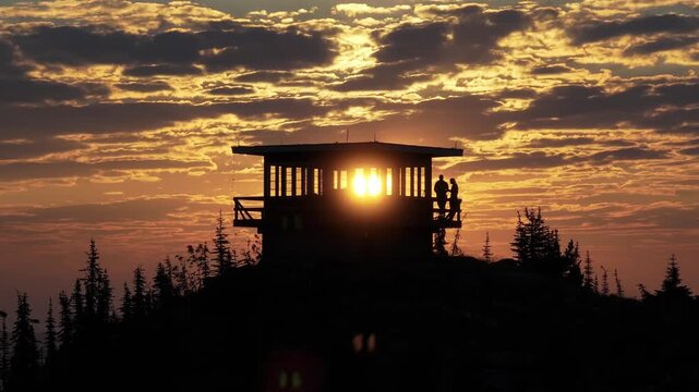 An aerial drone video of a couple standing on the porch balcony of a fire lookout tower during an epic gorgeous sunrise as the sun is revealed through the tower windows taken with a DJI Mavic 3 Pro