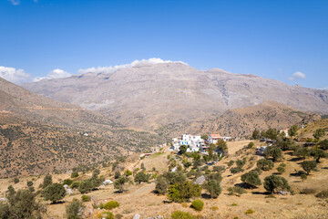 Small cluster of whitewashed buildings sits on a sunlit, dry hillside dotted with olive trees, set against expansive rugged mountains under a clear blue sky in southern Crete. The landscape is open