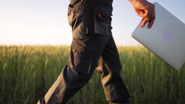 Walking with laptop through field farmer in workwear carries device by hand across crop at sunset showcasing agriculture modern technology in rural landscape and computer use for farm management