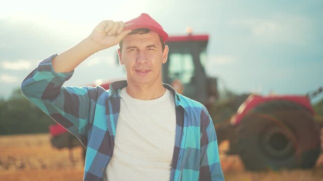 Farmer standing in field under sunlight near tractor wearing red hat and plaid shirt inspecting crop preparing for harvest on rural farm as worker handling soil and equipment tire and plow visible