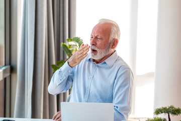 Tired senior businessman yawning while working on laptop at office. Exhausted elderly male...