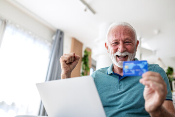 Happy senior man holding credit card using laptop at home. Excited elderly male buyer making online...