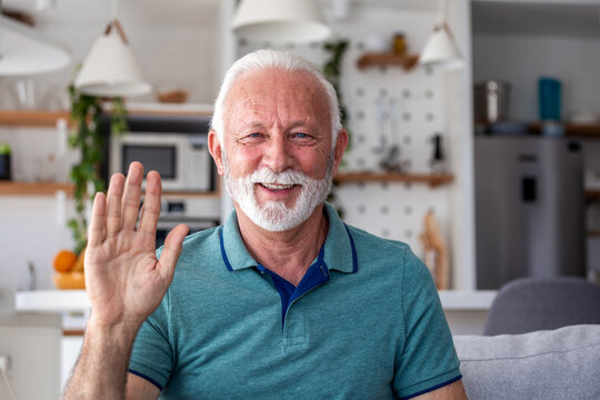 Happy senior man waving hand at camera during video call at home. Friendly elderly male greeting family or doctor through webcam. Communication, retirement lifestyle and technology concept.