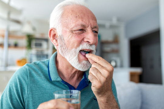 Taking medication. Senior man putting a pill in his mouth and holding a glass of water. Elderly male patient taking supplements or prescription medicine, healthcare and medicine concept at home.