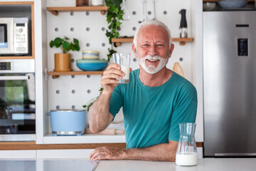 Portrait of happy senior man holding glass of milk in kitchen. Elderly male in green shirt smiling at camera, retirement lifestyle. Grandpa enjoying healthy beverage and nutrition at home indoors.