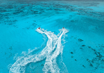 Fototapeta premium Aerial view of jet skis over shallow turquoise sea with reef in Zanzibar. Clear tropical water, watercraft trails, summer vacation, coastal travel and marine leisure activity. Top drone view. Exotic