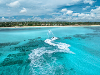 Fototapeta premium Aerial view of jet skis over shallow turquoise sea, sandy beach, palm trees in Zanzibar. Clear tropical water, watercraft trails, summer vacation, coastal travel and marine leisure activity. Top view
